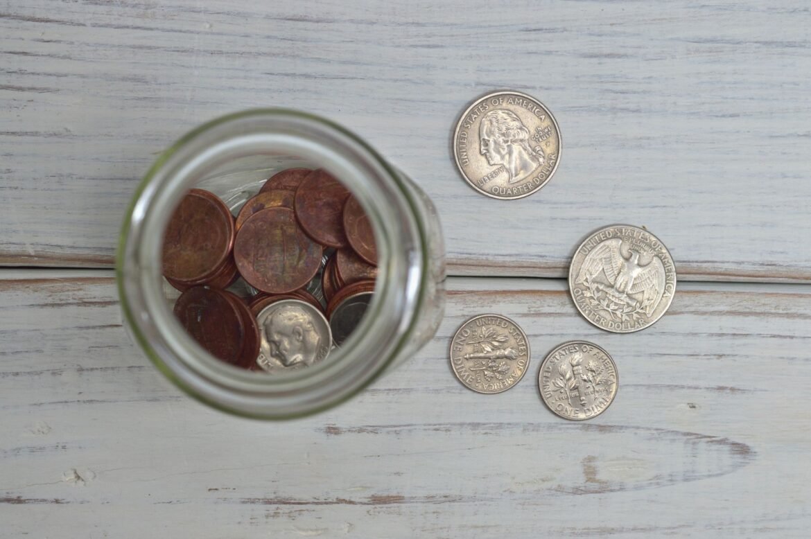 A glass jar filled with coins standing on a wooden table.