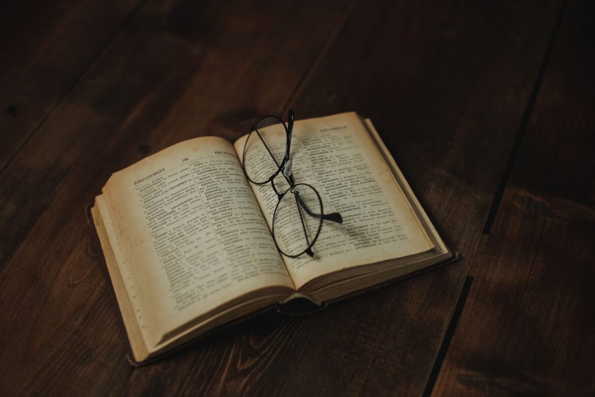 An open vintage book on a wooden surface with a pair of eyeglasses resting on the page.
