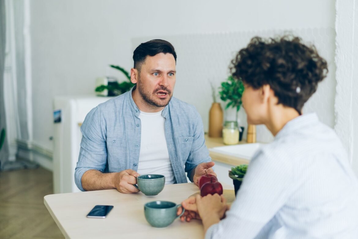 A couple sitting across from each other at a kitchen table with coffee cups, mid-conversation.