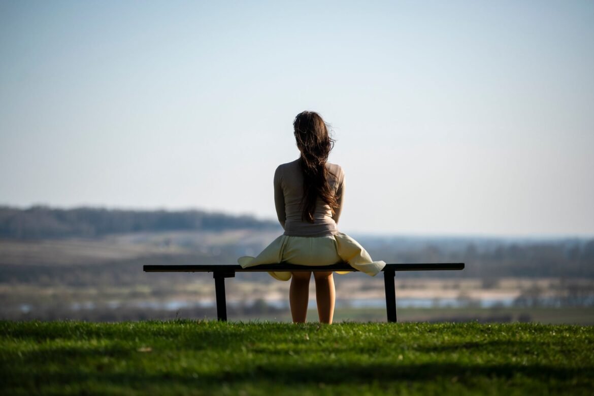 A woman sits alone on a bench looking out over an open field.