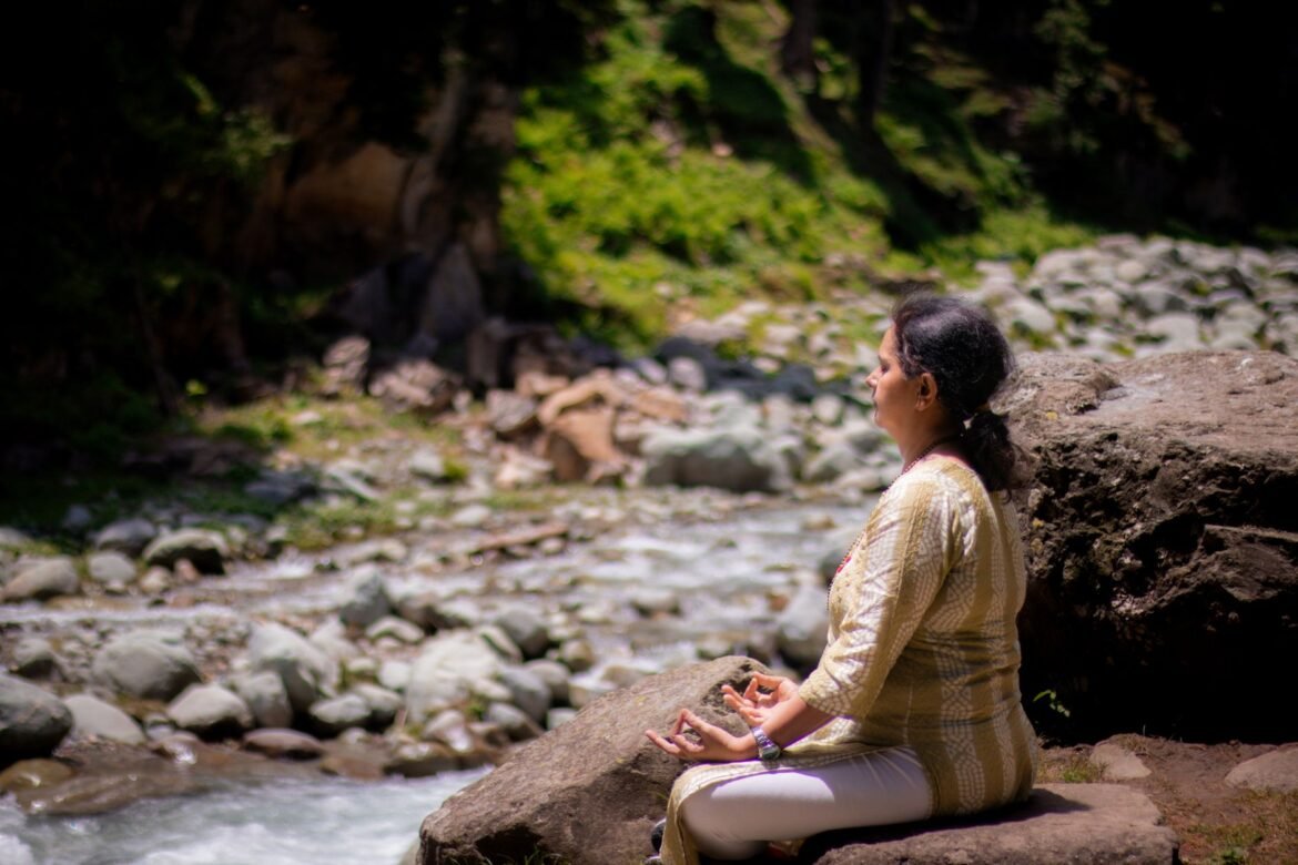 A woman sitting in meditation on rocks beside a river, surrounded by greenery.