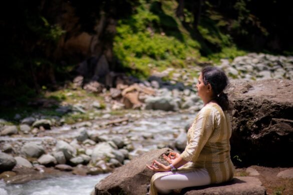 A woman sitting in meditation on rocks beside a river, surrounded by greenery.