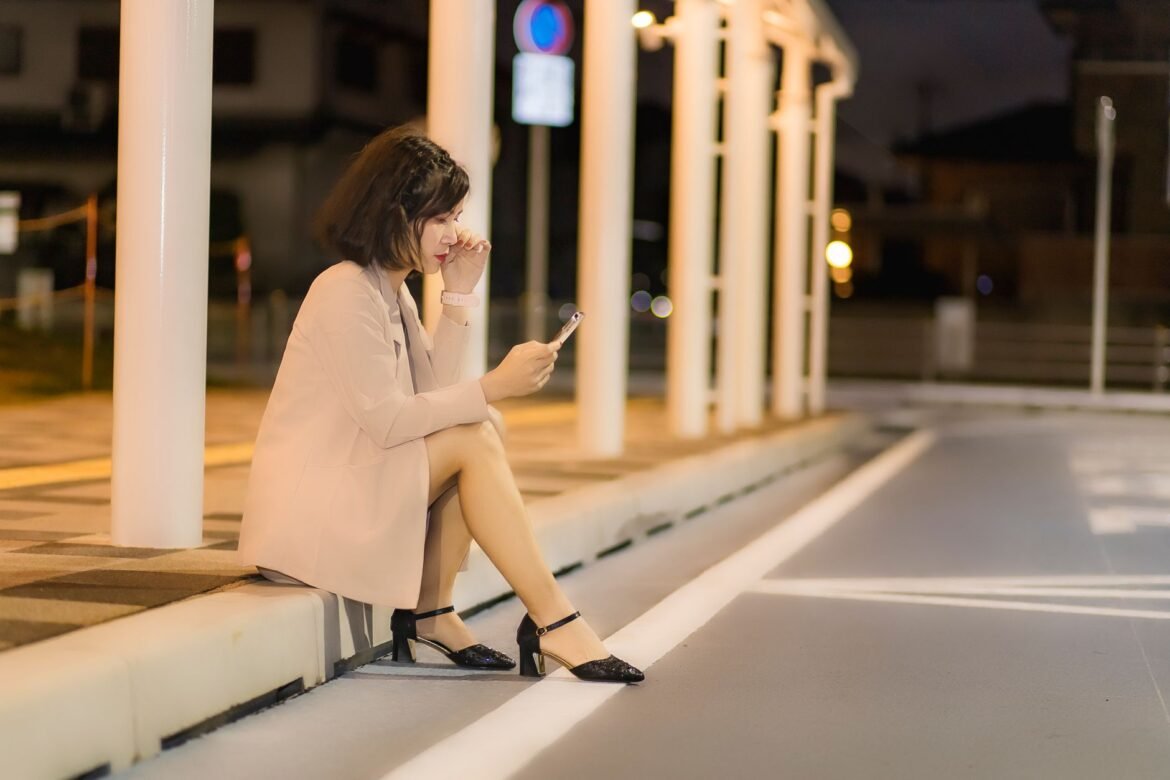 A woman sitting on a sidewalk at night, looking down at her phone.