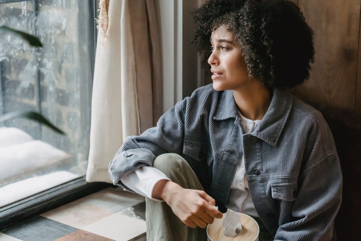 A woman sits on a windowsill with her head lowered, looking away from the camera.
