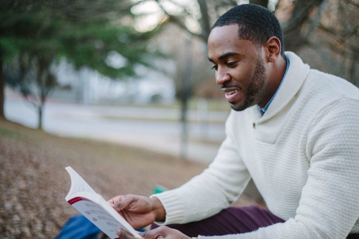 A young man sitting outdoors in a park, reading a book.