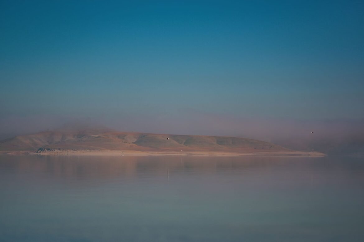 Calm water reflecting mountains at dawn, low mist across the surface